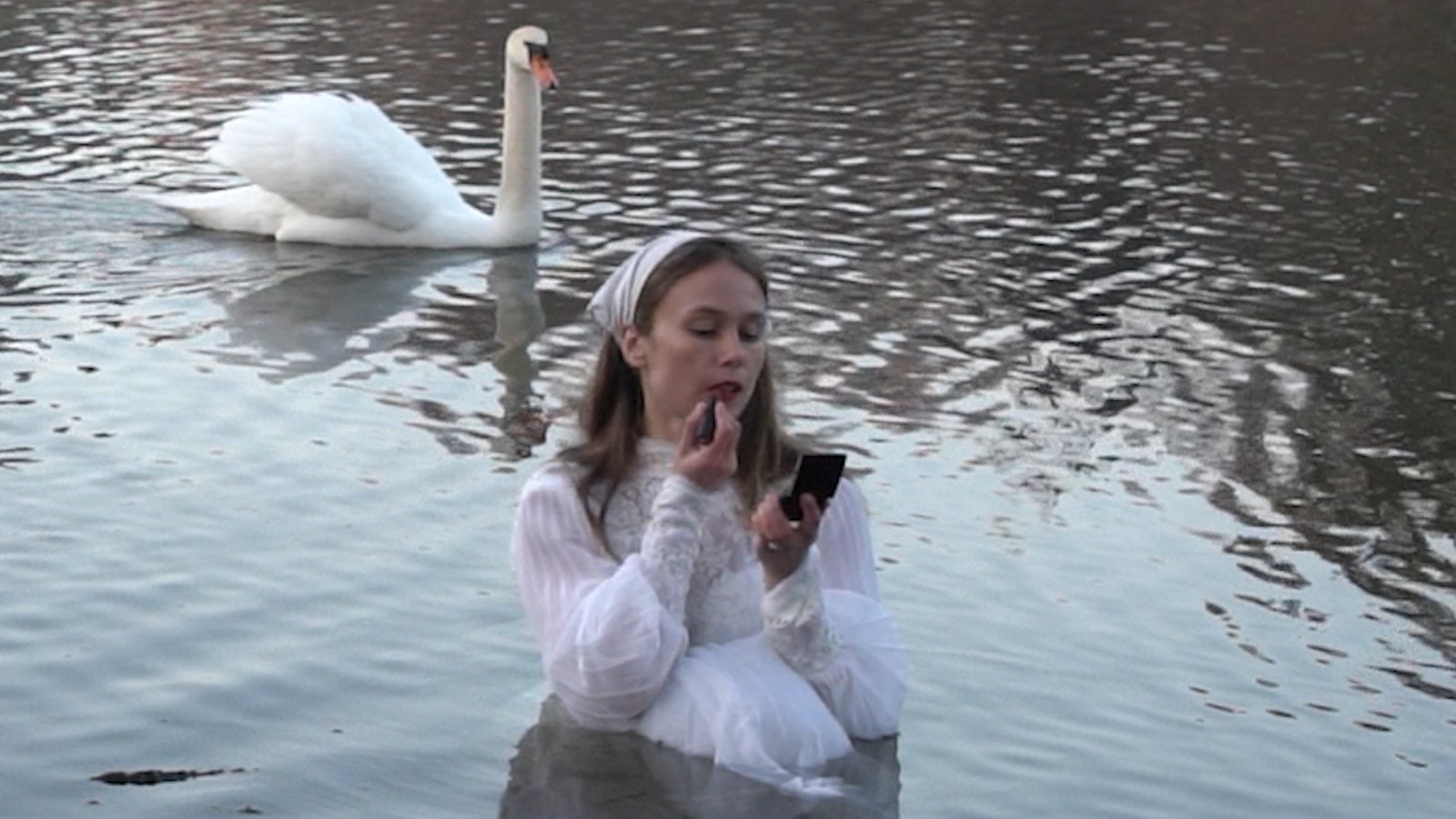 Photograph by artist Sage Szkabarnicki-Stuart, of herself standing in a lake, wearing a white dress and applying lipstick, with a white swan in the background