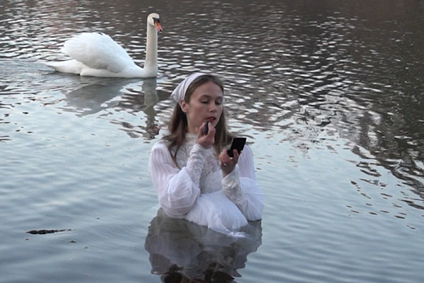 Photograph by artist Sage Szkabarnicki-Stuart, of herself standing in a lake, wearing a white dress and applying lipstick, with a white swan in the background