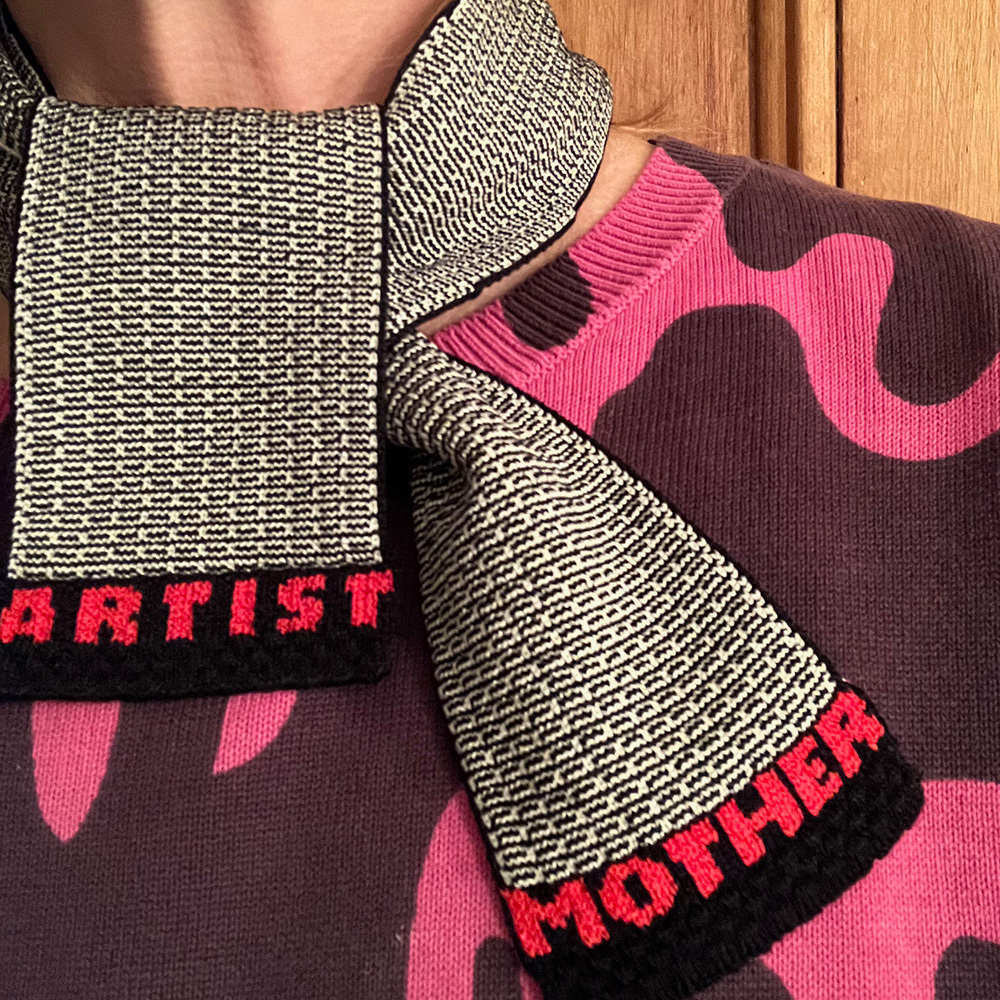 Close-up of a torso wearing a pink and purple sweater and a scarf that reads 