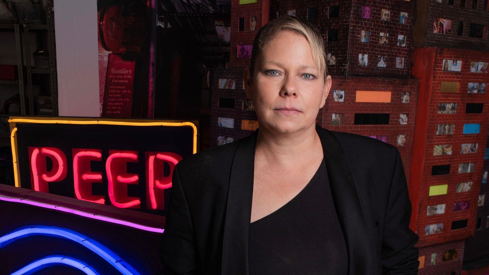 Artist Tracey Snelling stands in front of her exhibition, featuring a neon light and a building installation in the background.