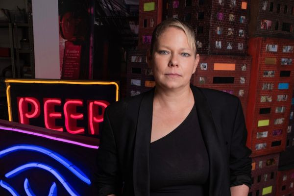 Artist Tracey Snelling stands in front of her exhibition, featuring a neon light and a building installation in the background.